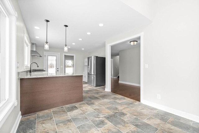 kitchen featuring wall chimney range hood, sink, hanging light fixtures, stainless steel refrigerator, and light stone counters