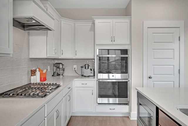 kitchen featuring white cabinetry, light hardwood / wood-style flooring, backsplash, custom exhaust hood, and appliances with stainless steel finishes