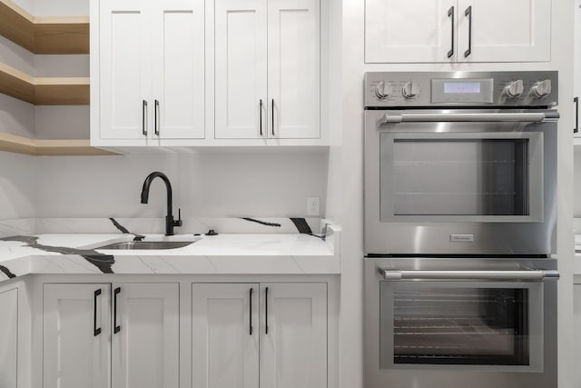 kitchen featuring white cabinets, stainless steel double oven, light stone counters, and sink