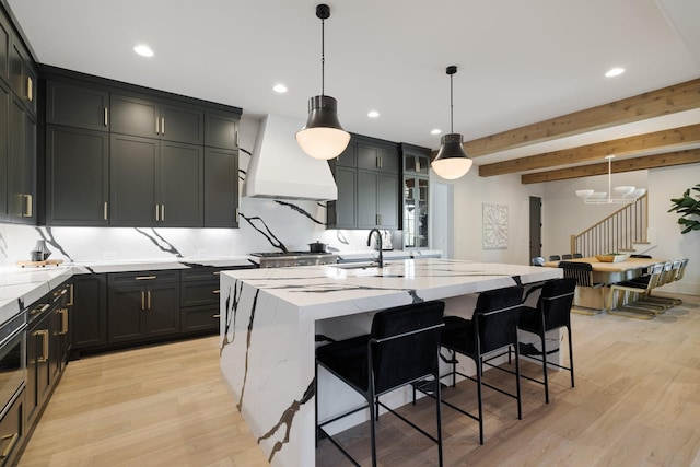 kitchen featuring pendant lighting, premium range hood, a center island with sink, light stone countertops, and beamed ceiling