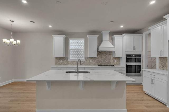 kitchen with custom exhaust hood, an island with sink, sink, and light stone counters