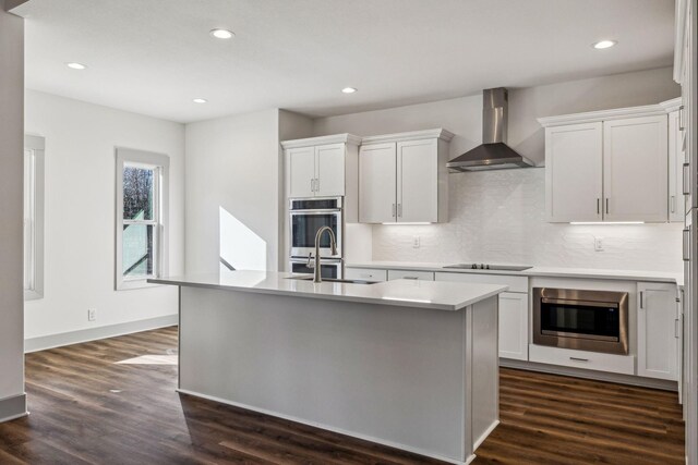 kitchen featuring wall chimney exhaust hood, dark wood-type flooring, a kitchen island with sink, white cabinets, and appliances with stainless steel finishes