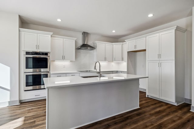 kitchen featuring white cabinets, stainless steel double oven, wall chimney exhaust hood, and an island with sink