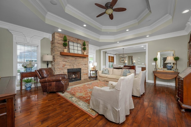 living room with ceiling fan with notable chandelier, a tray ceiling, a stone fireplace, and ornamental molding