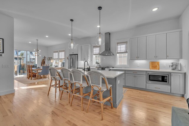 kitchen with pendant lighting, a breakfast bar, a center island with sink, wall chimney range hood, and stainless steel appliances