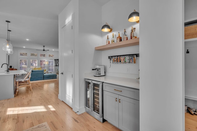 bar with gray cabinetry, ceiling fan, hanging light fixtures, beverage cooler, and light hardwood / wood-style floors