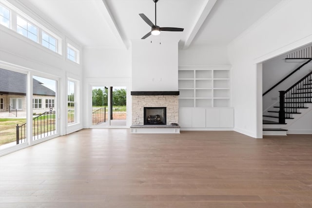 unfurnished living room with built in shelves, ceiling fan, a stone fireplace, beamed ceiling, and hardwood / wood-style flooring