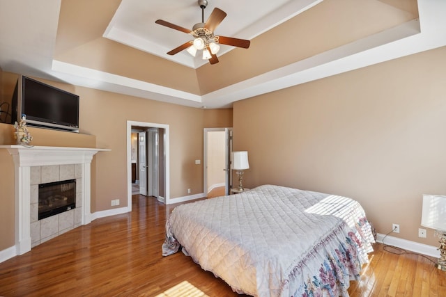 bedroom with ceiling fan, a tiled fireplace, a raised ceiling, and wood-type flooring