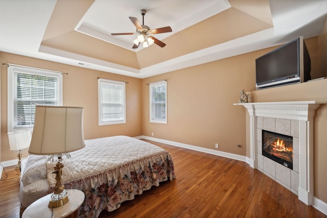 bedroom with a tile fireplace, ceiling fan, a tray ceiling, and hardwood / wood-style floors