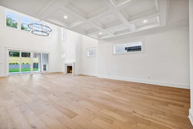 unfurnished living room featuring coffered ceiling, light wood-type flooring, a towering ceiling, beam ceiling, and an inviting chandelier