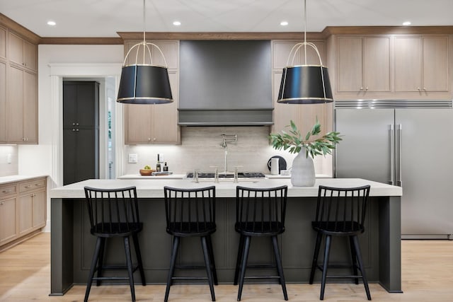 kitchen featuring an island with sink, built in refrigerator, hanging light fixtures, and light brown cabinets