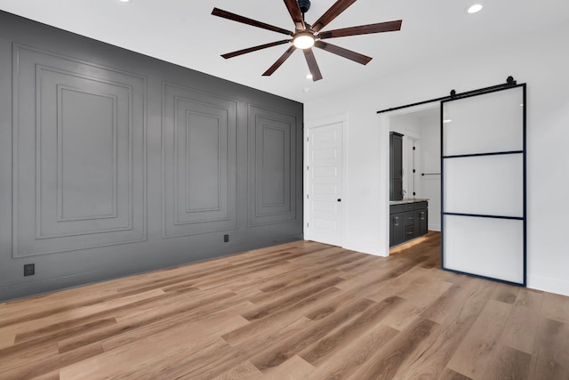 unfurnished bedroom featuring connected bathroom, ceiling fan, a barn door, and light hardwood / wood-style flooring