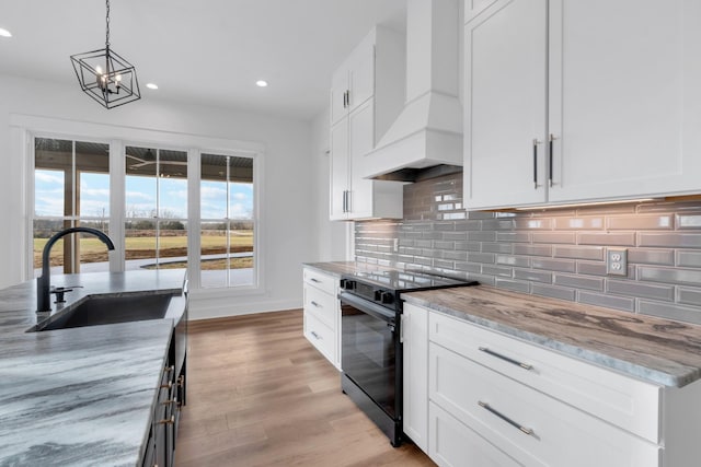 kitchen with light stone countertops, custom range hood, white cabinetry, black electric range, and sink