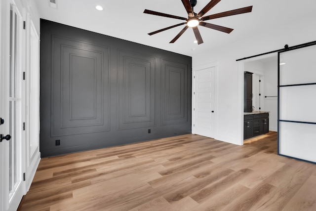 unfurnished bedroom featuring ceiling fan, light hardwood / wood-style flooring, connected bathroom, and a barn door