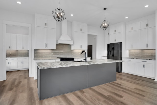 kitchen featuring black fridge with ice dispenser, pendant lighting, an island with sink, custom range hood, and white cabinetry