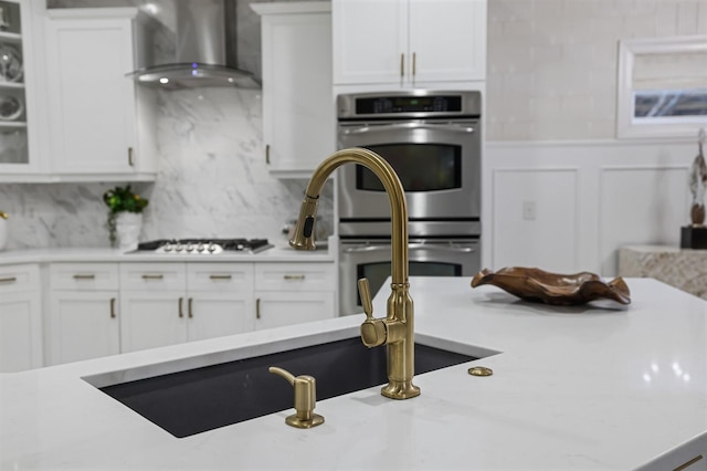 kitchen with gas stovetop, stainless steel double oven, decorative backsplash, white cabinetry, and wall chimney range hood