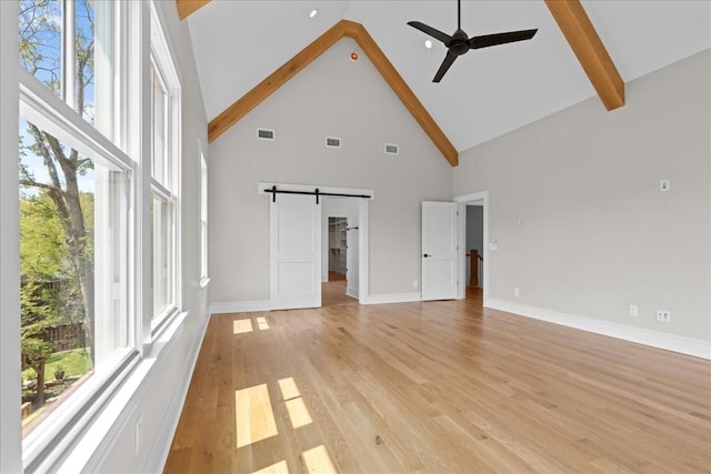 unfurnished living room featuring ceiling fan, light wood-type flooring, a barn door, high vaulted ceiling, and beam ceiling