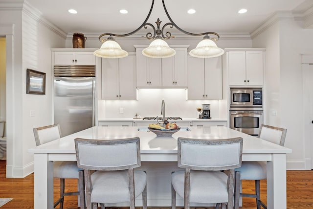 kitchen featuring built in appliances, a kitchen island with sink, pendant lighting, and white cabinetry