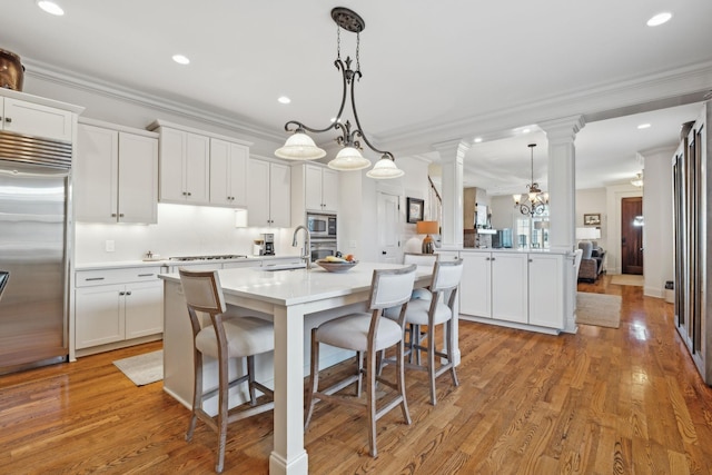 kitchen featuring an island with sink, built in appliances, a notable chandelier, white cabinetry, and decorative light fixtures