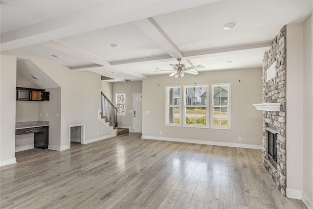 unfurnished living room with ceiling fan, beamed ceiling, a stone fireplace, and light hardwood / wood-style flooring