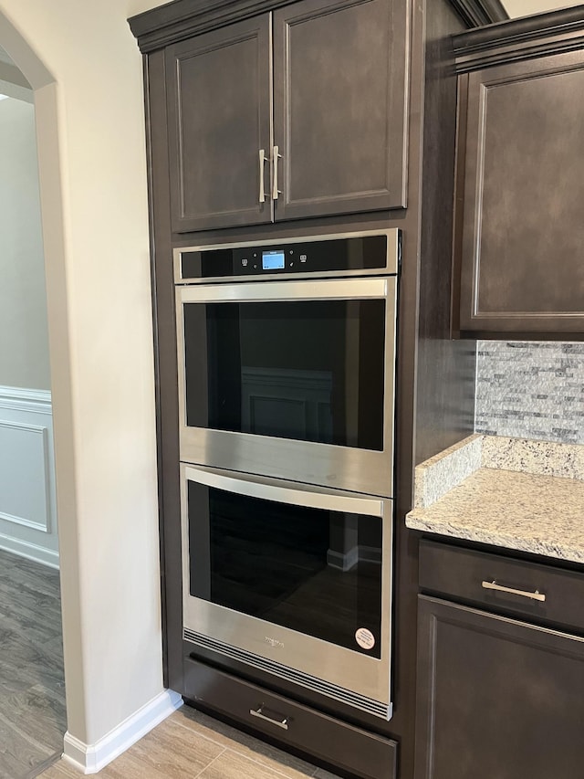 kitchen featuring light stone counters, light wood-type flooring, backsplash, stainless steel double oven, and dark brown cabinetry