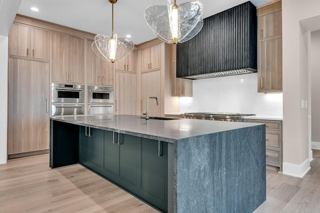 kitchen with premium range hood, hanging light fixtures, an island with sink, light brown cabinetry, and sink