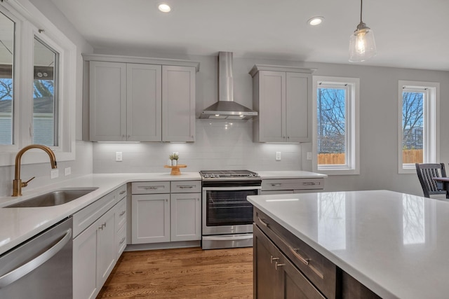 kitchen with wall chimney exhaust hood, stainless steel appliances, tasteful backsplash, sink, and hanging light fixtures