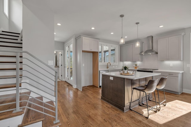 kitchen with a kitchen island, wall chimney range hood, hanging light fixtures, light wood-type flooring, and stainless steel dishwasher