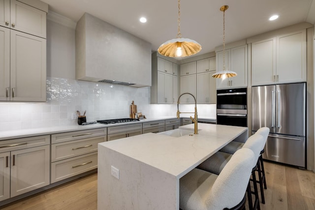 kitchen featuring appliances with stainless steel finishes, an island with sink, sink, wall chimney range hood, and backsplash