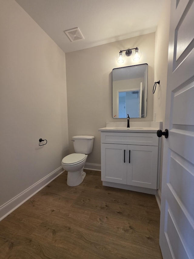 half bath featuring vanity, visible vents, toilet, wood finished floors, and baseboards