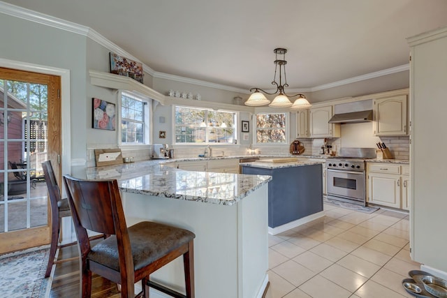 kitchen featuring wall chimney exhaust hood, high end range, decorative light fixtures, backsplash, and light tile patterned flooring