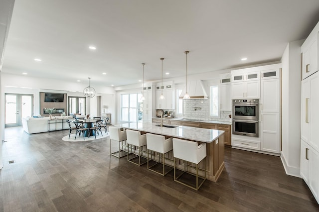 kitchen with premium range hood, a breakfast bar, a center island with sink, stainless steel double oven, and white cabinets