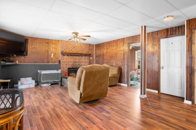 living room featuring wood walls, a brick fireplace, ceiling fan, hardwood / wood-style floors, and a drop ceiling