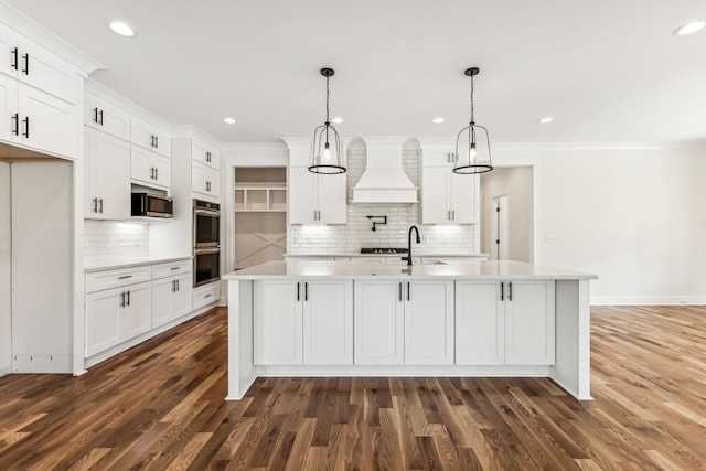 kitchen with decorative light fixtures, a kitchen island with sink, premium range hood, and appliances with stainless steel finishes