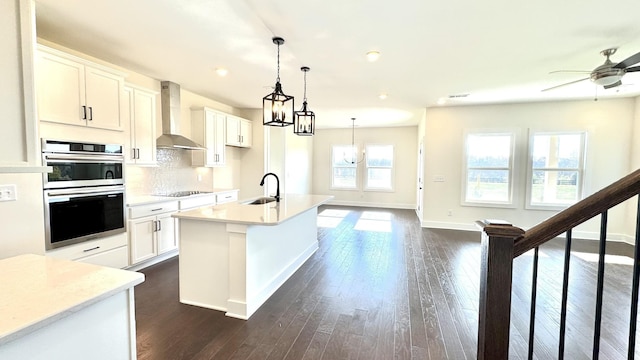 kitchen with double oven, white cabinetry, wall chimney range hood, and an island with sink