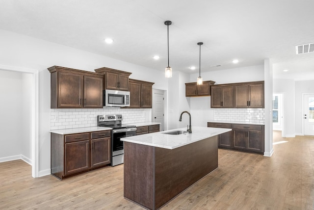 kitchen with sink, appliances with stainless steel finishes, pendant lighting, light stone countertops, and a kitchen island with sink