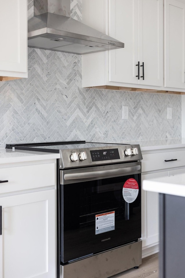 kitchen with light wood-type flooring, stainless steel range with electric stovetop, decorative backsplash, wall chimney exhaust hood, and white cabinetry