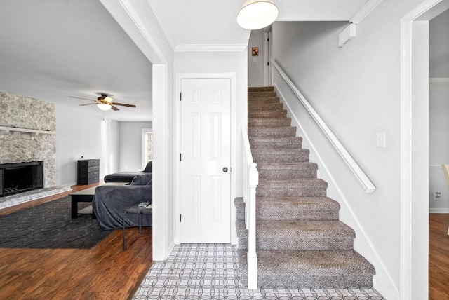 stairway featuring ceiling fan, wood-type flooring, a stone fireplace, and ornamental molding