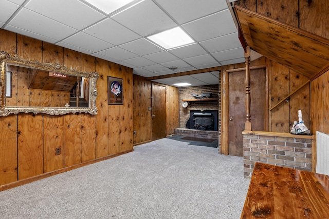 unfurnished living room featuring light colored carpet, a drop ceiling, wooden walls, a wood stove, and bar area