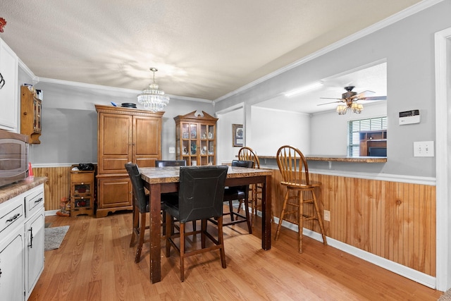 dining space with ceiling fan with notable chandelier, a textured ceiling, wood walls, ornamental molding, and light wood-type flooring