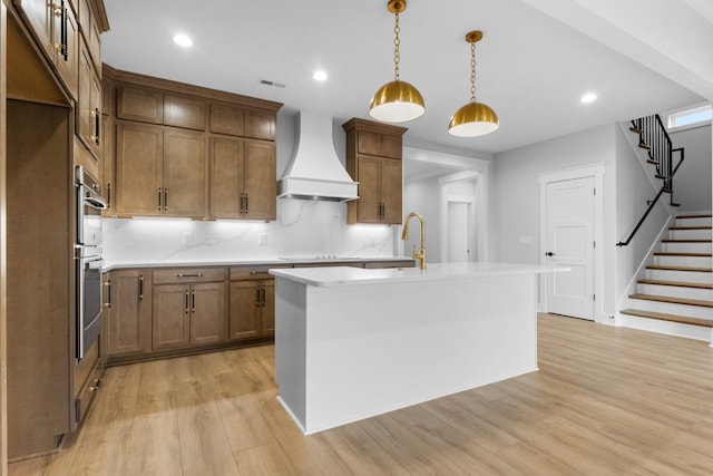 kitchen with custom range hood, light wood-type flooring, backsplash, hanging light fixtures, and a kitchen island with sink