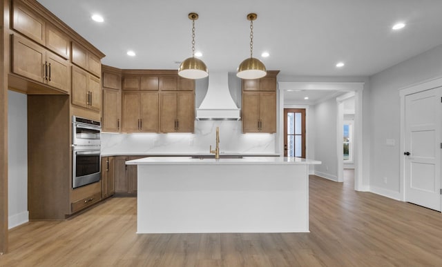 kitchen featuring stainless steel double oven, an island with sink, custom range hood, and hanging light fixtures