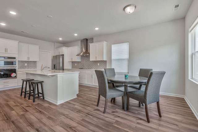 kitchen featuring white cabinets, a kitchen bar, a center island with sink, wall chimney range hood, and appliances with stainless steel finishes