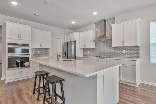 kitchen featuring wall chimney exhaust hood, stainless steel appliances, a kitchen island with sink, backsplash, and white cabinets