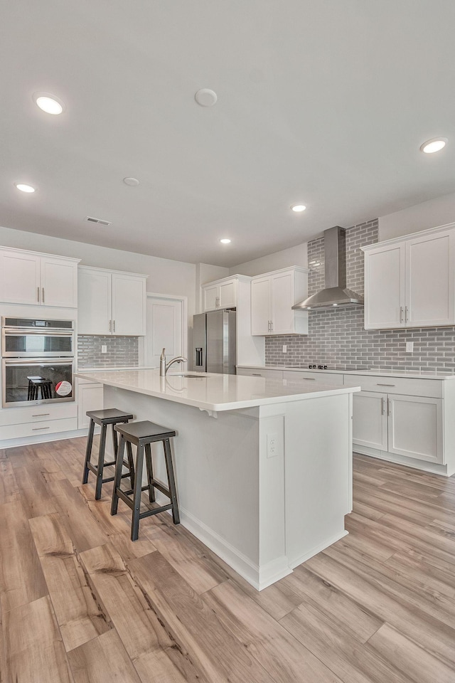 kitchen featuring white cabinets, a kitchen island with sink, wall chimney exhaust hood, and stainless steel appliances