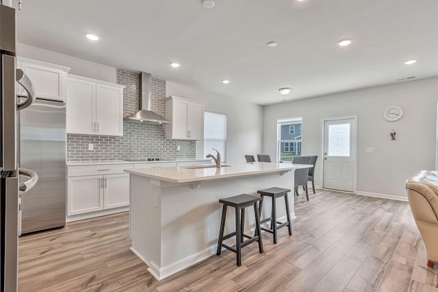 kitchen with decorative backsplash, wall chimney range hood, a kitchen island with sink, and white cabinetry