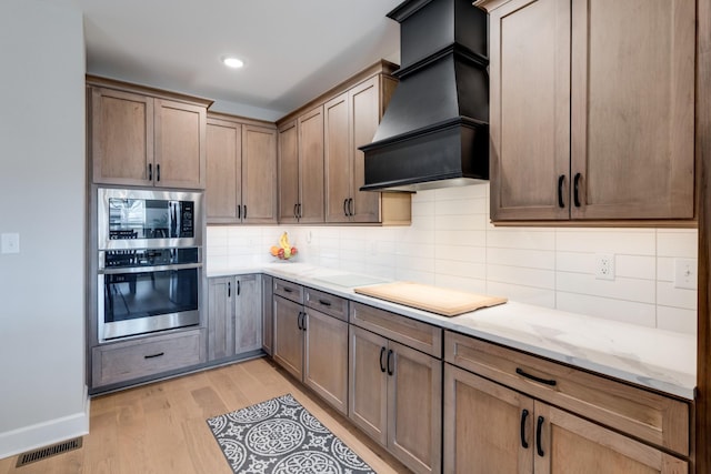 kitchen with tasteful backsplash, light hardwood / wood-style floors, appliances with stainless steel finishes, custom range hood, and light stone counters