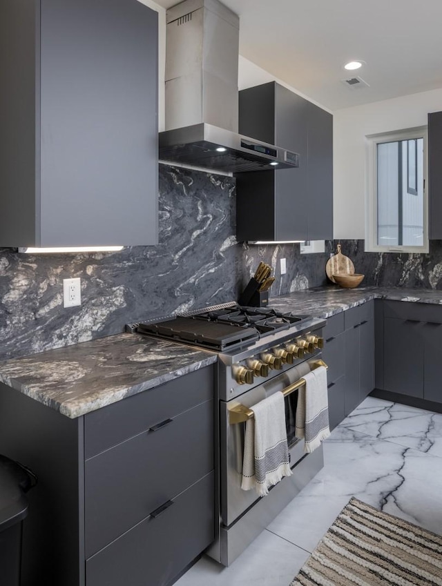 kitchen featuring dark stone countertops, gray cabinets, stainless steel gas range oven, wall chimney range hood, and backsplash