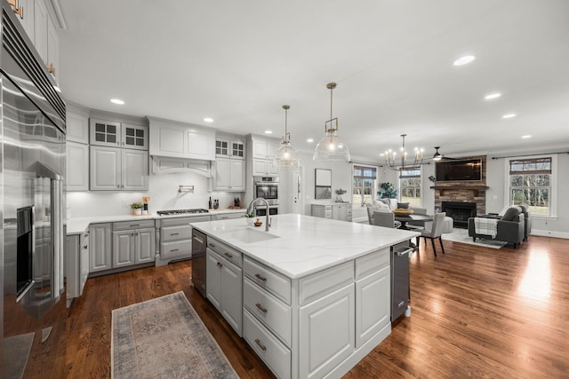 kitchen featuring light stone countertops, a stone fireplace, an island with sink, ceiling fan, and sink