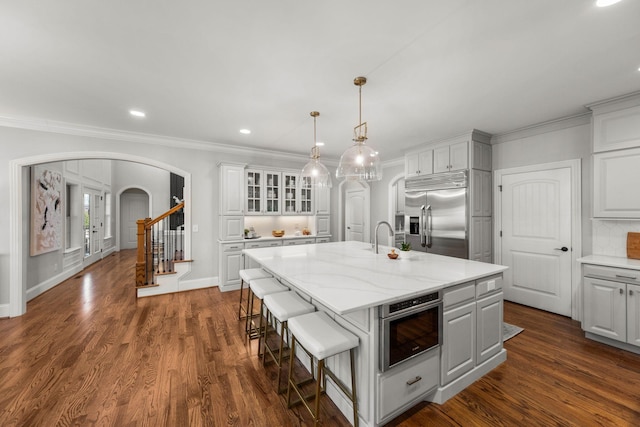 kitchen with hanging light fixtures, an island with sink, stainless steel built in refrigerator, light stone counters, and white cabinetry